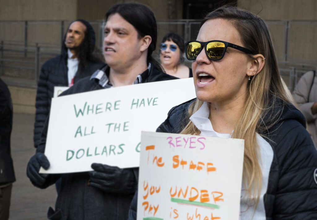 people holding signs, one which says "where have all the dollars gone" and anothe rwhich says "reyes - the staff you under pay is what... [cut off]"