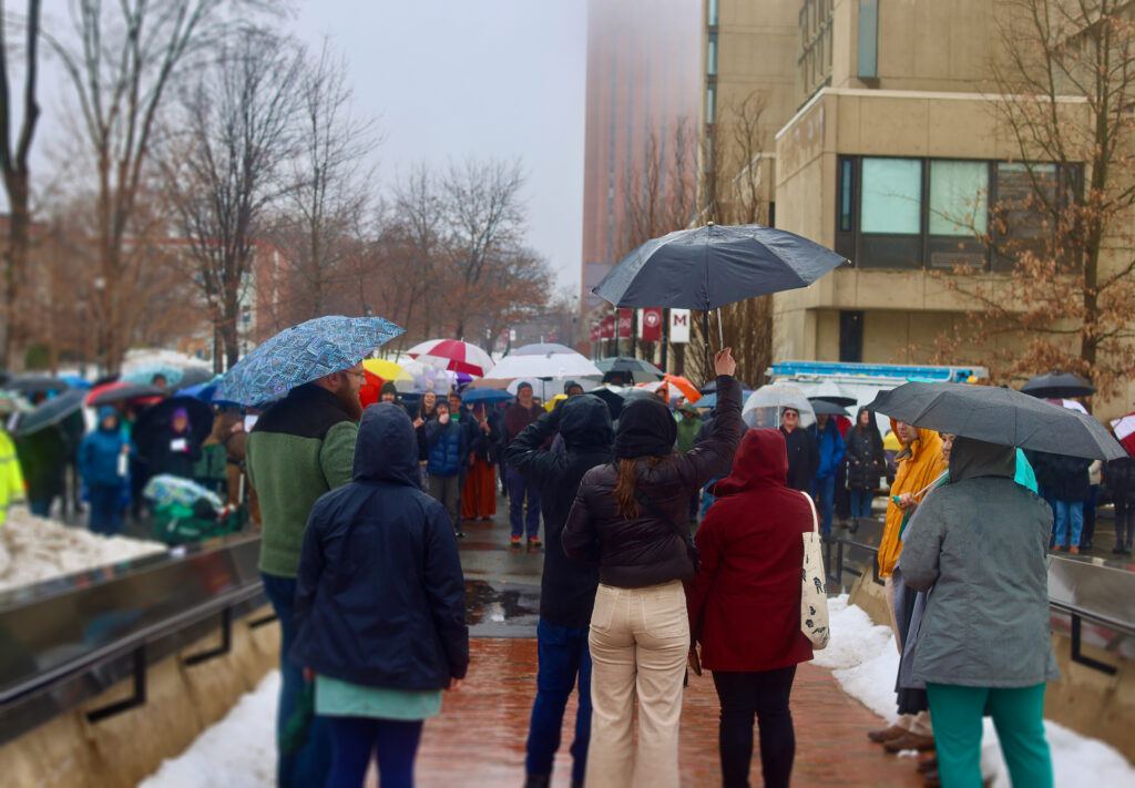 People standing out to make their voices heard near the Whitmore administrative building on a rainy snowy day, holding umbrellas and standing together. 